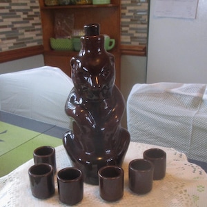 May include: A brown ceramic squirrel-shaped decanter with a matching stopper, accompanied by six small shot glasses. The set is displayed on a white lace tablecloth. The decanter is approximately 10 inches tall.