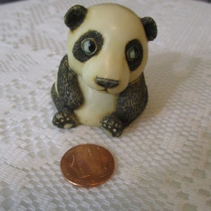May include: A small, white ceramic panda figurine with black ears, eyes, and paws. The panda is sitting with its paws tucked in front of it. The figurine is about the size of a US penny.