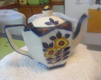 May include: A vintage porcelain teapot with a white base, adorned with navy blue accents and floral designs in yellow and orange. The teapot features a gold-colored handle, spout, and lid finial, and is set on a white lace tablecloth.