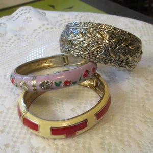May include: Three hinged bangle bracelets. The top bracelet is silver-toned metal with an embossed leaf pattern. The middle bracelet is light purple enamel with small red and green floral accents. The bottom bracelet is cream enamel with red rectangular inlays.