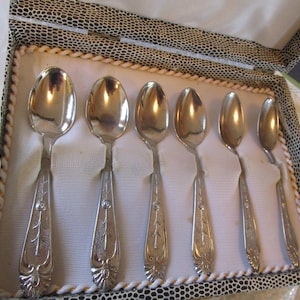 May include: A set of six silver spoons displayed in a decorative box. The spoons have ornate handles with a floral design. The box has a black and white spotted pattern on the exterior and a white interior.
