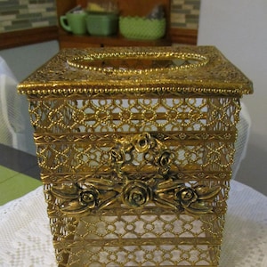May include: A square gold-tone metal tissue box cover featuring an intricate openwork lattice design. The front panel is decorated with a prominent three-dimensional floral motif consisting of sculpted roses and leaves centered on the mesh surface.