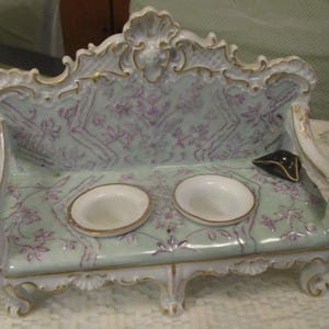 May include: A decorative porcelain sofa-shaped dish with two small bowls. The sofa is painted in pale green and white with gold trim and purple floral accents. A small black hat sits on the sofa.