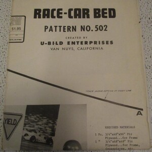 May include: Vintage "Race-Car Bed" pattern booklet. The cover features bold black text on a cream background, with the title and pattern number prominently displayed. Includes a black and white image of a race car and a yield sign. Created by U-Bild Enterprises.