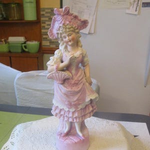 May include: A porcelain figurine of a woman in a pink dress and hat, holding a basket. The dress has white lace trim and ruffles. The figurine is standing on a pink base. The woman has blonde hair and is smiling.