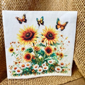 May include: A square paper napkin featuring a printed illustration of two large sunflowers, several smaller sunflowers, and a cluster of white daisies at the base. Three orange and yellow butterflies are positioned above the flowers.