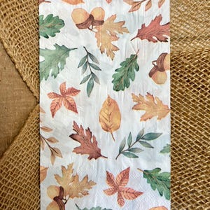 May include: White paper napkin with a watercolor design of autumn leaves and acorns in shades of green, orange, and brown. The napkin is rectangular and features a repeating pattern of fall foliage.