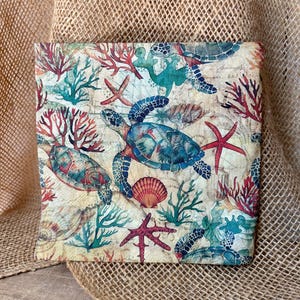 May include: A square fabric item with a sea turtle design. The pattern includes sea turtles, starfish, coral, and seashells in shades of blue, red, and green on a cream-colored background. The item is likely a decorative pillow or fabric panel.