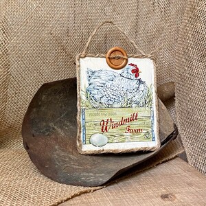 May include: A rustic wooden sign with a white chicken and a white egg in a crate. The sign says "Fresh Eggs" and "Windmill Farm".