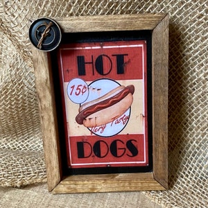 May include: A vintage-style wooden sign with a red, white, and black design. The sign reads "HOT 15¢ Very Tasty DOGS". A cartoon hot dog with mustard and ketchup is pictured on the sign. The sign is framed in a rustic brown wood frame.