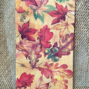 May include: A rectangular paper napkin featuring a fall-themed design. The napkin is covered with detailed illustrations of autumn leaves in shades of red, orange, yellow, and green, along with acorns. The background is a light beige.