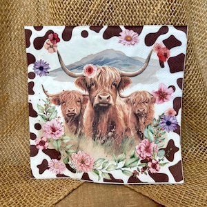 May include: Square decorative napkin featuring a watercolor illustration of three Highland cows with long horns, surrounded by pink and purple flowers. The napkin has a white background with brown cow spots and a mountain in the background.