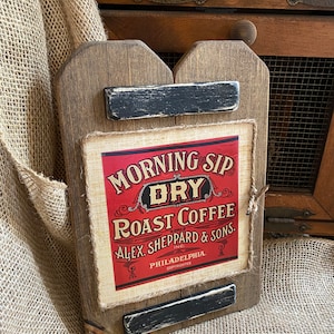 May include: A rustic wooden sign with a distressed finish. The sign features a rectangular label with the text "MORNING SIP DRY ROAST COFFEE" in a vintage style. The label is red and cream-colored. The sign is trimmed with black accents and twine.
