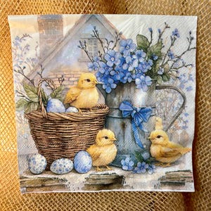 May include: A decorative paper napkin featuring an Easter theme. The design includes a woven basket with blue speckled eggs and yellow chicks. A metal pitcher with blue flowers and a blue bow is also present, along with a small house in the background.