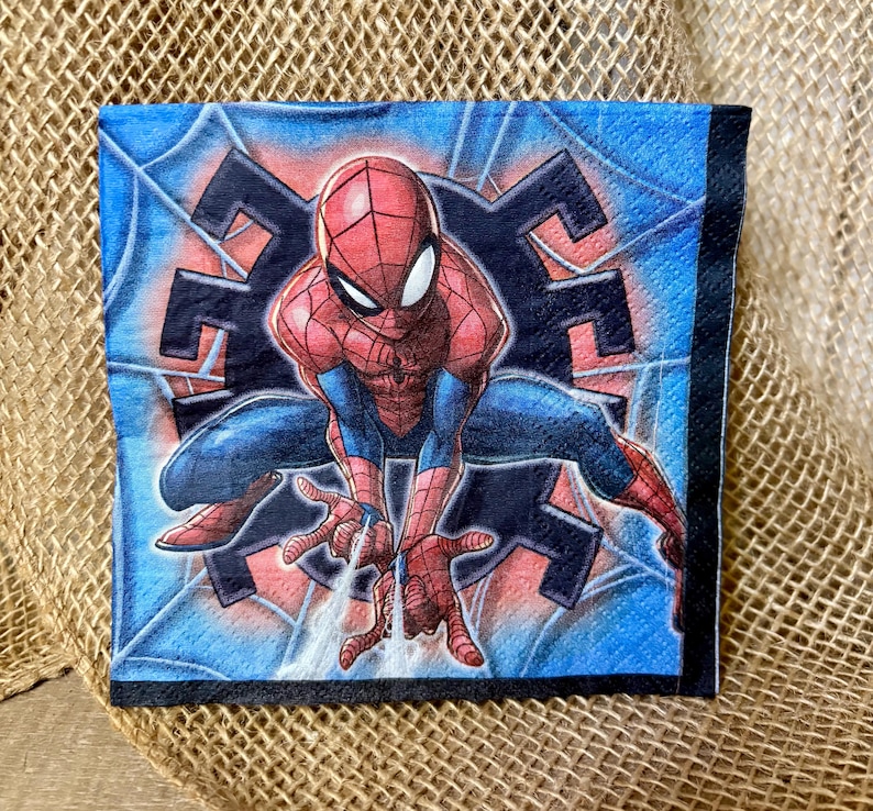 May include: A square paper napkin featuring a graphic illustration of the superhero Spider-Man in a crouching pose. The character is red and blue with a black web pattern, set against a blue background with a dark spider web design.