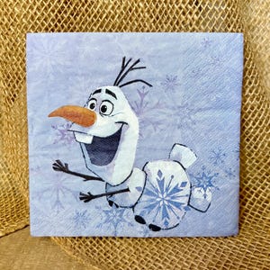 May include: A square paper napkin featuring a light blue background with a pattern of white and blue snowflakes. The central graphic depicts a white snowman character with a large orange carrot nose, stick arms, and a wide, happy expression.
