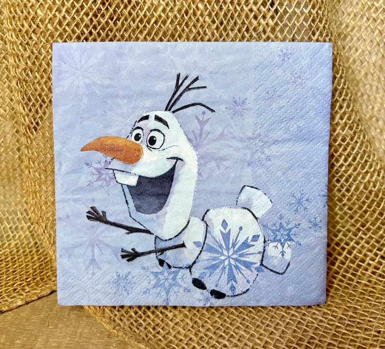 May include: A square paper napkin featuring a light blue background with a pattern of white and blue snowflakes. The central graphic depicts a white snowman character with a large orange carrot nose, stick arms, and a wide, happy expression.