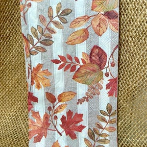 May include: A folded rectangular napkin with a fall leaves pattern. The leaves are in shades of red, orange, and brown, set against a background of vertical gray and white stripes. The napkin is likely made of paper or fabric.
