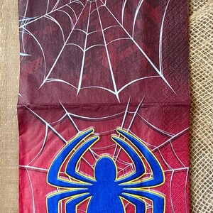Puede incluir: Servilleta de papel roja y blanca con un dise&ntilde;o de telara&ntilde;a azul y amarilla. La telara&ntilde;a tiene la forma de una ara&ntilde;a. El texto "MARVEL" est&aacute; impreso en la servilleta.