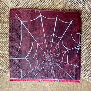 Puede incluir: Servilleta de papel roja con un dise&ntilde;o de telara&ntilde;a blanca. El texto "Hallmark Cards Inc." est&aacute; impreso en la servilleta.