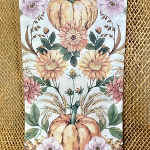 May include: A rectangular paper napkin featuring a symmetrical botanical print with two orange pumpkins, light pink and peach flowers, green leaves, and wheat stalks on a white background. The word Unique is printed in small text on the left side.