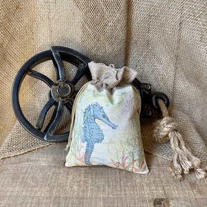 May include: A small burlap bag with a drawstring closure, featuring a printed design of a blue seahorse and coral. The bag is sitting on a rustic wooden surface with a black metal wheel in the background.
