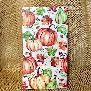 May include: A rectangular paper napkin featuring a watercolor-style print of orange, white, and green pumpkins interspersed with colorful autumn leaves, small berries, and winding vines against a clean white background.