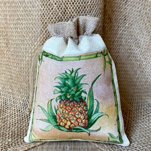 May include: A small burlap drawstring bag with a white fabric lining and a printed image of a pineapple on a light pink background. The pineapple is surrounded by green leaves and a bamboo border.