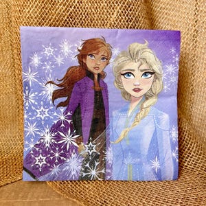 May include: A square paper napkin featuring a purple background with white snowflake patterns. The illustration depicts two characters: one with long auburn hair wearing a purple coat, and one with long blonde hair in a braid wearing a blue coat.