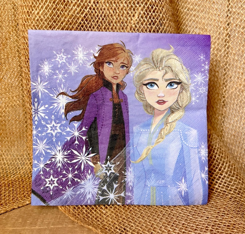 May include: A square paper napkin featuring a purple background with white snowflake patterns. The illustration depicts two characters: one with long auburn hair wearing a purple coat, and one with long blonde hair in a braid wearing a blue coat.