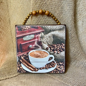 May include: A wooden sign with a rustic design featuring a cup of coffee, cinnamon sticks, and coffee beans. The sign has a wooden bead handle.