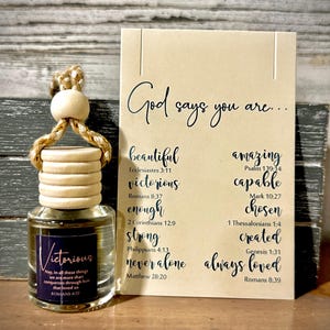 May include: An air freshener with a wooden top and a glass bottle labeled "Victorious." A card next to it reads "God says you are..." with words like "beautiful" and "amazing." The air freshener is approximately 3 inches tall.