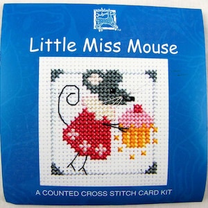 Peut inclure: Un kit de point de croix compté représentant une souris de dessin animé portant une robe rouge à pois blancs et tenant un cupcake rose et jaune. Le kit est intitulé "Little Miss Mouse".