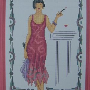 Peut inclure: Un motif de point de croix représentant une femme en robe rose debout à côté d'une table avec un verre de liquide rouge dessus. La femme tient une cigarette dans sa main droite et un éventail dans sa main gauche. L'arrière-plan est un cadre blanc avec une bordure verte et violette.