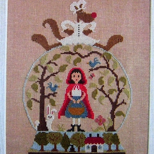 May include: Cross-stitch embroidery of a snow globe with a red-hooded girl, a wolf in a dress, and a forest scene. The snow globe is on a base with a small house and trees.