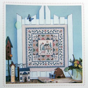 May include: A cross-stitch sampler featuring a house with a red roof, surrounded by trees and flowers. The sampler is framed by a white picket fence and a blue butterfly is flying above. The sampler is displayed on a table with other decorative items, including a birdhouse, a watering can, and a beehive.