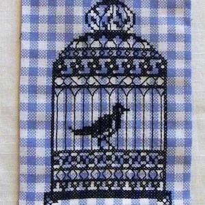 Puede incluir: Una jaula de pájaros bordada en blanco y negro con un pájaro negro en su interior, colgada de un colgador dorado con una campana. El bordado está sobre una tela de cuadros azules y blancos.