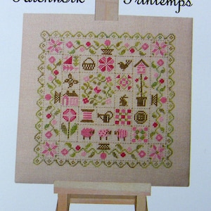 Peut inclure: Un sampler de point de croix sur un chevalet en bois. Le sampler est beige avec un motif floral vert et rose et les mots "Patchwork Printemps".