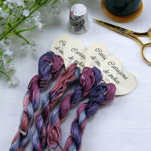 May include: Four skeins of embroidery floss in shades of purple, pink, and blue, each tied with a heart-shaped tag that reads "Cartagena de Indias".