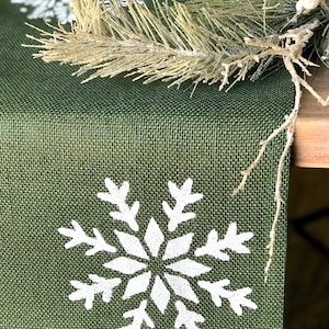 May include: A green burlap table runner with a white snowflake design. The runner is decorated with a sprig of faux pine.