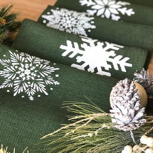 May include: A set of green burlap table runners with white snowflake designs. The runners are arranged in a stack, with the top runner showing a large snowflake design. The runners are decorated with pine sprigs and a pine cone.