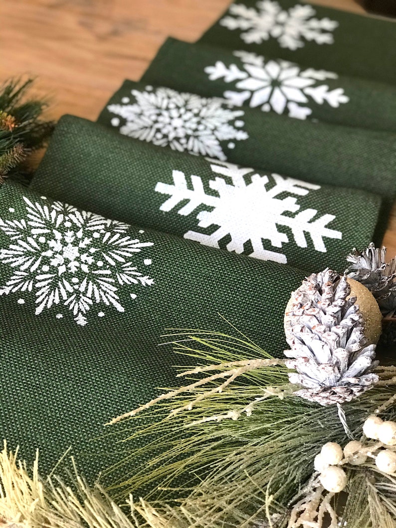 May include: A set of green burlap table runners with white snowflake designs. The runners are arranged in a stack, with the top runner showing a large snowflake design. The runners are decorated with pine sprigs and a pine cone.