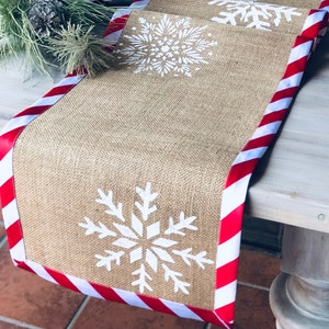 May include: A burlap table runner with a red and white striped border. The runner features three white snowflake designs.