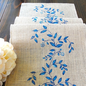 May include: Set of four burlap placemats with a blue floral design. The placemats are made of natural burlap fabric and feature a repeating pattern of blue leaves and flowers.