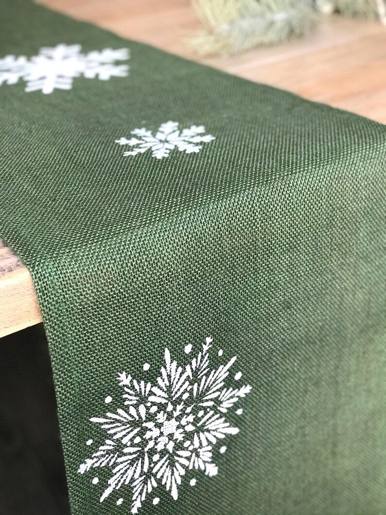May include: A green burlap table runner with white snowflake designs.