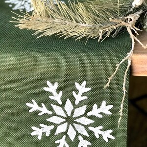 May include: A green burlap table runner with white snowflake designs. The runner is decorated with a sprig of faux pine.