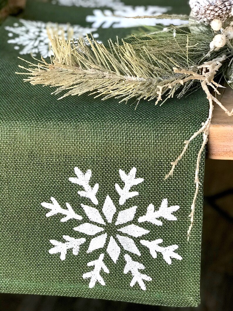 May include: A green burlap table runner with white snowflake designs. The runner is decorated with a sprig of faux pine.