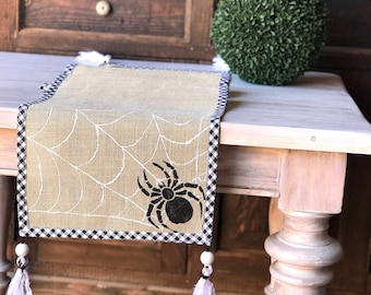May include: A rectangular burlap table runner featuring a white printed spiderweb pattern and a black spider graphic. The runner is bordered with a black and white checkered fabric trim and finished with white wooden beads and white fabric tassels.