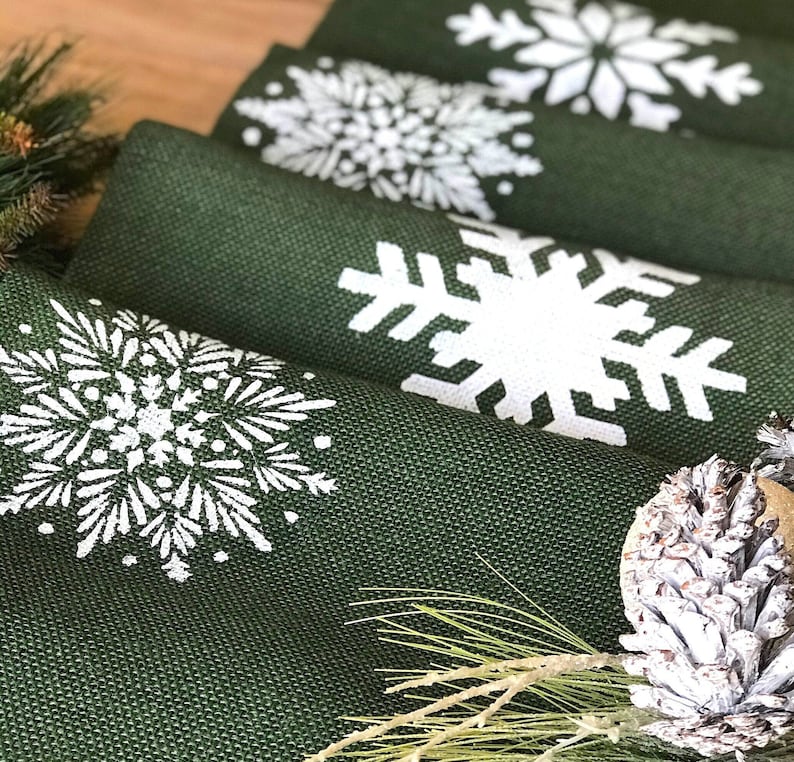 May include: A set of four green burlap placemats with white snowflake designs. The placemats are folded and stacked on top of each other.