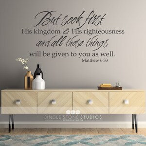 May include: A wall decal with the Bible verse "But seek first His kingdom & His righteousness and all these things will be given to you as well. Matthew 6:33". The decal is on a gray wall above a wooden dresser with three drawers. There are three vases on the dresser, one gold, one black, and one white.